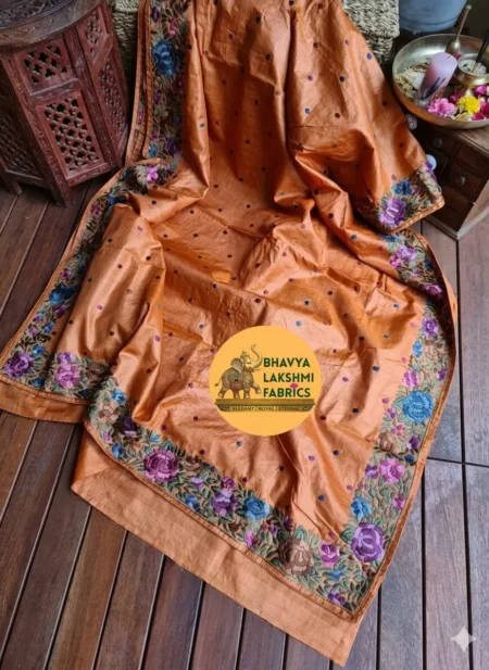 A luxurious rust orange pure Tussar Munga silk saree laid flat, featuring scattered small dark embroidered dots across the body and an intricate border adorned with vibrant multicolored floral embroidery in shades of purple, violet, blue, and green leaves. The textile displays a rich, textured wild silk grain
