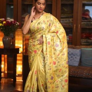 A model poses elegantly in a luxurious sunshine yellow pure Tussar Munga Bhagalpuri silk saree. The saree is adorned with intricate all-over floral jaal embroidery featuring pink, violet, and orange blossoms on delicate green vines. The palla features neat hand-knotted tassels.