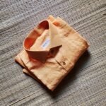 A luxurious peach orange pure handloom linen shirt laid flat on a woven straw mat background. The shirt features a clean button-down collar, a neat front chest pocket, and a white brand label reading Bhavya Loom
