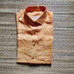 A luxurious peach orange pure handloom linen shirt laid flat on a woven straw mat background. The shirt features a clean button-down collar, a neat front chest pocket, and a white brand label reading Bhavya Loom