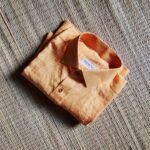 A luxurious peach orange pure handloom linen shirt laid flat on a woven straw mat background. The shirt features a clean button-down collar, a neat front chest pocket, and a white brand label reading Bhavya Loom
