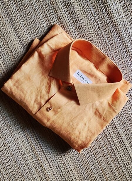 A luxurious peach orange pure handloom linen shirt laid flat on a woven straw mat background. The shirt features a clean button-down collar, a neat front chest pocket, and a white brand label reading Bhavya Loom